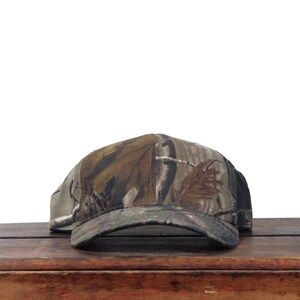 May include: A baseball cap featuring a realistic forest camouflage pattern in shades of brown, tan, and dark green. The cap has a structured crown and a curved brim, with the camouflage print extending across the entire front and side panels.