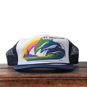 May include: A navy blue and white trucker hat featuring a colorful graphic of dolphins jumping over waves under a rainbow. The text "ST. AUGUSTINE" is printed above the graphic. The hat has a mesh back.
