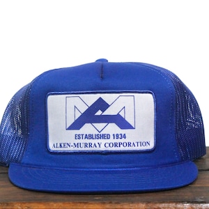 May include: A royal blue trucker hat with a white mesh back. The hat has a blue patch with the text "ESTABLISHED 1934 ALKEN-MURRAY CORPORATION" and a stylised "MA" logo.