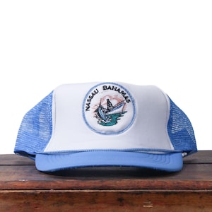 Könnte beinhalten: Weiß-blaue Trucker-Cap mit gebogenem Schirm. Die Kappe hat einen runden Aufnäher mit den Worten "Nassau Bahamas" und einem Bootsbild. Die Rückseite der Kappe ist aus blauem Mesh.