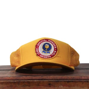Peut inclure: Casquette de camionneur jaune avec un dos en filet et une visière incurvée. Le chapeau présente un écusson circulaire avec le texte "OFFICIAL MEMBER" et "PABST DRINKING TEAM" autour d'un emblème bleu et or.