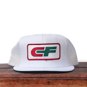 May include: White trucker hat with a red and green CF logo patch. The hat has a white front panel, a curved brim, and a mesh back. The logo is in a rectangular shape.