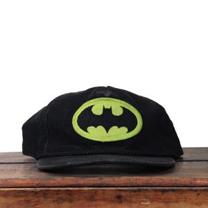 May include: Black corduroy baseball cap with a neon green Batman logo. The cap has a curved peak and a classic design. The Batman logo is an oval with a bat symbol in the centre.