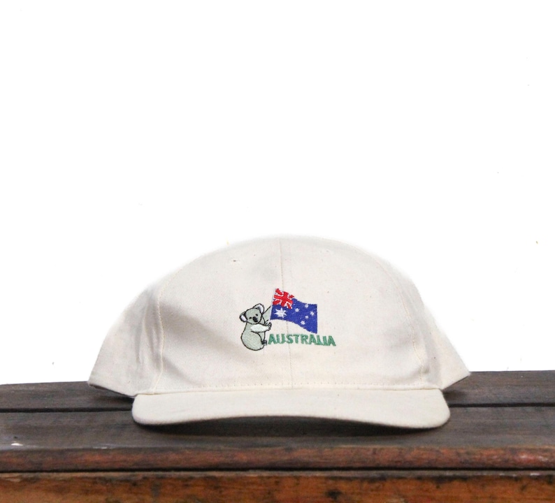 May include: A white baseball cap with an embroidered koala holding an Australian flag and the word "AUSTRALIA" underneath.