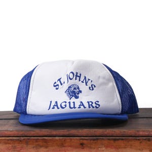 May include: A white and blue trucker hat with the text "ST. JOHN'S JAGUARS" in blue. The hat features a blue brim and mesh back, with a jaguar graphic. This vintage style hat is a great accessory.