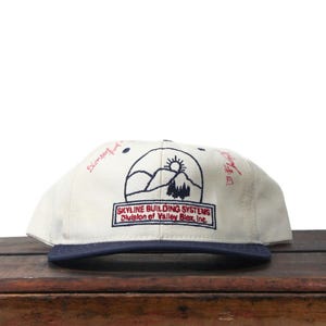 May include: A cream-coloured baseball cap with a navy blue brim. The cap features an embroidered logo with mountains, a sun, and text that reads "SKYLINE BUILDING SYSTEMS Division of Valley Blz, Inc.". Handwritten text in red is on the side.