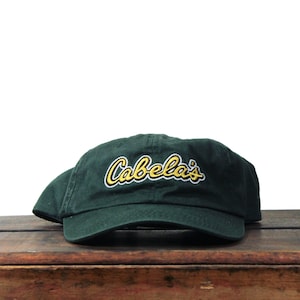 May include: A dark forest green baseball cap featuring the word Cabela's embroidered in yellow thread with a white outline across the front panel. The cap has a curved brim and a structured crown, showcasing a classic six-panel design with stitching.