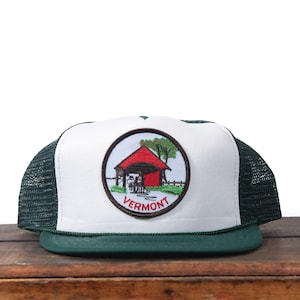 Könnte beinhalten: Weiß-grüne Trucker-Mütze mit rundem Vermont-Aufnäher. Der Aufnäher zeigt eine rote Scheune, Bäume und das Wort "Vermont". Die Mütze hat eine weiße Vorderseite, einen grünen Schirm und eine grüne Mesh-Rückseite.