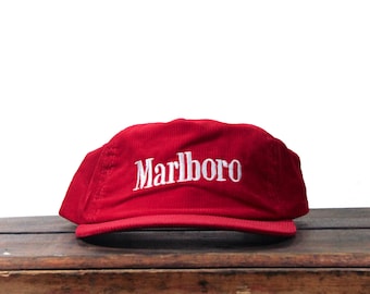 May include: A bright red corduroy baseball cap featuring the word Marlboro embroidered in white thread across the front panel. The hat has a structured crown and a curved brim, showcasing a textured fabric finish throughout the entire design.