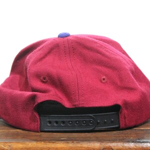May include: A maroon baseball cap with a black strap and a small blue patch on the top.