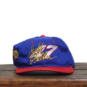 May include: Blue and red baseball cap with a white and gold embroidered logo that says "Jeff Gordon 24" on the front.
