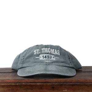 May include: A gray baseball cap with the text "ST. THOMAS" embroidered in white. Below the text is "1493" in a rectangular box. The cap is sitting on a wooden surface.