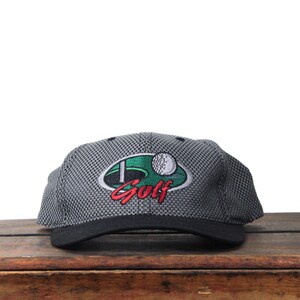Puede incluir: Gorra de golf gris y negra con visera negra. La gorra presenta un diseño bordado con la palabra "Golf" en rojo, un óvalo verde y una pelota de golf blanca.