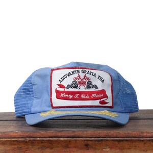 May include: A light blue trucker hat with a mesh back and a curved brim. The front patch features text and illustrations, including the words "ADJUVANTE GRATIA TUA" and "Henry J. Cole Shows". The brim has gold embroidery.