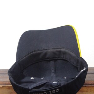 May include: Black baseball cap with a yellow stripe along the brim. The interior is black with a black sweatband. The text "cat.com" is visible on the front of the cap.