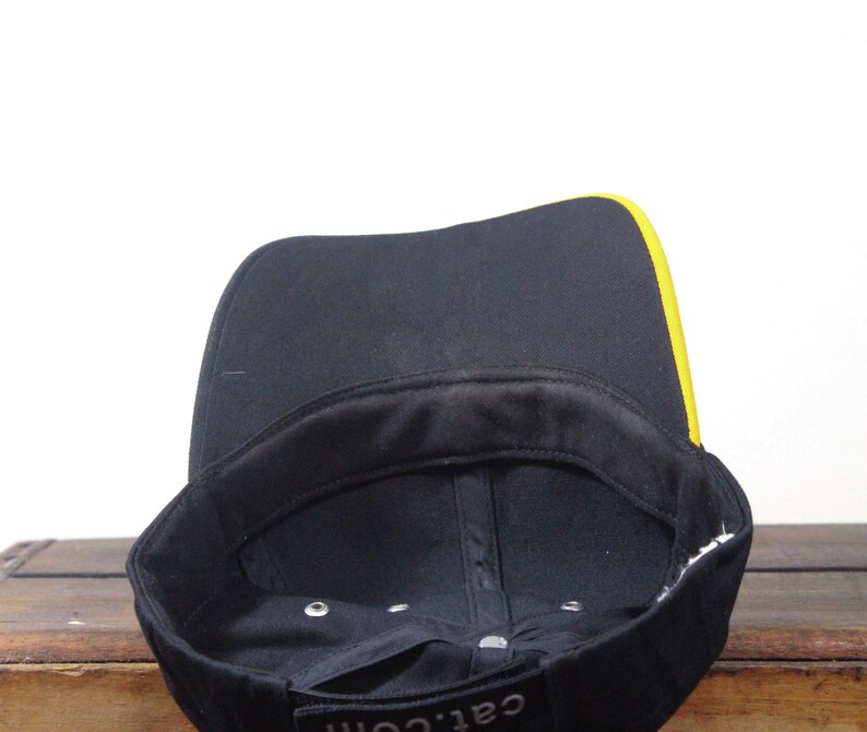 May include: Black baseball cap with a yellow stripe along the brim. The interior is black with a black sweatband. The text "cat.com" is visible on the front of the cap.