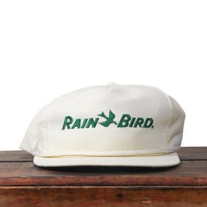 Pode incluir: Boné de beisebol creme com as palavras "RAIN BIRD" bordadas em verde, juntamente com um gráfico de pássaro. O boné tem uma aba plana e um detalhe de corda na base. Está sobre uma superfície de madeira.