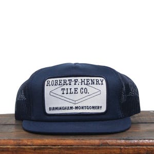 May include: A navy blue trucker hat with a white patch that reads "Robert F. Henry Tile Co. Birmingham-Montgomery".