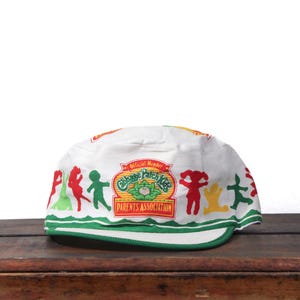 May include: White Cabbage Patch Kids Parents Association cap with a green brim. The cap features colorful cartoon figures and the Cabbage Patch Kids logo in red, green, and yellow. The text "Official Member" is also visible.
