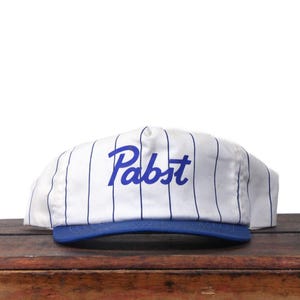 Puede incluir: Gorra de béisbol a rayas blancas y azules con la palabra "Pabst" en azul en la parte delantera. La visera es azul. La gorra está sobre una superficie de madera.