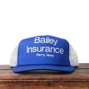 May include: A blue and white trucker hat with the text "Bailey Insurance Perry, Iowa" in white. The hat has a blue front panel and a white mesh back.