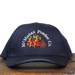 Puede incluir: Una gorra de béisbol azul marino con un logotipo bordado que dice "McMahan Powder Co." y presenta una imagen estilizada de un fuego artificial explotando.