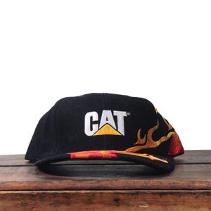 Puede incluir: Gorra de b&eacute;isbol negra con el logo CAT en blanco y amarillo. La gorra presenta un dise&ntilde;o de llama bordada en tonos naranja, amarillo y rojo. El sombrero est&aacute; sobre una superficie de madera.