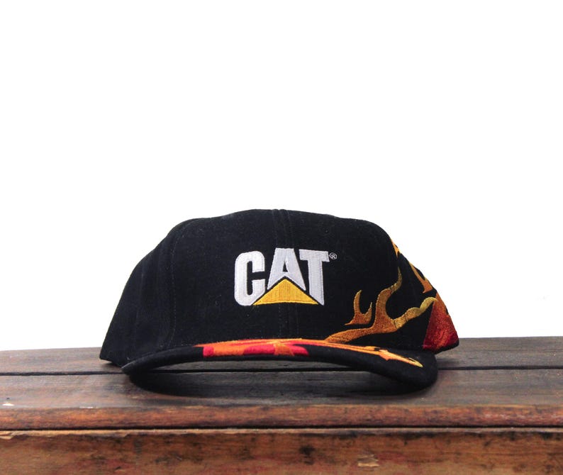 Puede incluir: Gorra de b&eacute;isbol negra con el logo CAT en blanco y amarillo. La gorra presenta un dise&ntilde;o de llama bordada en tonos naranja, amarillo y rojo. El sombrero est&aacute; sobre una superficie de madera.