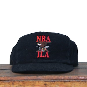 May include: Black corduroy baseball cap with the embroidered text "NRA ILA" in red, along with an eagle emblem. The cap has a flat brim and a structured crown.