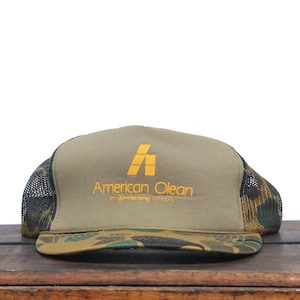 May include: A green, brown, and black camouflage trucker hat with a brown front panel. The front panel has a yellow logo that says "American Olean on Armstrong Carpets".