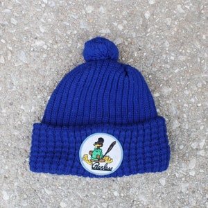 May include: Blue knitted beanie with a pom pom on top and a circular patch featuring a cartoon character holding a baseball bat. The patch says "Charles".