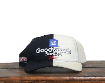 Könnte beinhalten: Eine schwarze und weiße Baseballkappe mit einem geteilten Design. Die Vorderseite der Kappe ist weiß mit dem Text "Goodwrench Service Plus" in schwarzer und blauer Schrift. Die Rückseite der Kappe ist schwarz mit einem kleinen blauen Quadrat mit den Buchstaben "GM" in weiß.