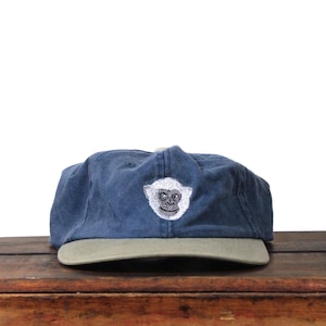 Peut inclure: Casquette de baseball bleu marine avec une visière vert kaki. La casquette présente un visage de singe blanc brodé sur le devant. Le chapeau est fait d'une matière douce et usée.