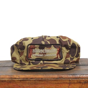 May include: A camouflage baseball cap with a brown patch that reads "Rough Country Chewing Tobacco" with a picture of a person fishing.