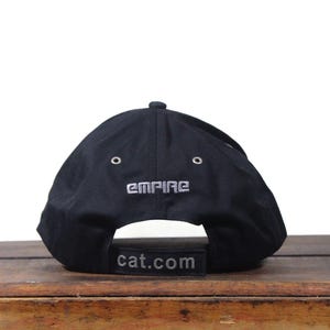 May include: Black baseball cap with the word "EMPIRE" embroidered in silver thread on the back. The curved brim features the text "cat.com" in a rectangular patch. The cap is sitting on a wooden surface.