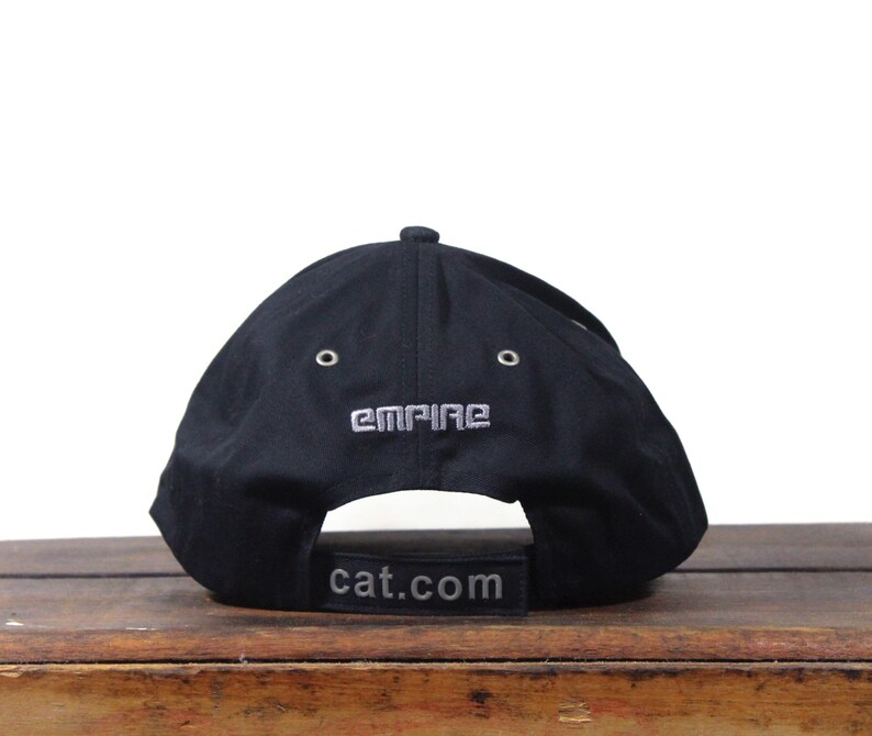May include: Black baseball cap with the word "EMPIRE" embroidered in silver thread on the back. The curved brim features the text "cat.com" in a rectangular patch. The cap is sitting on a wooden surface.