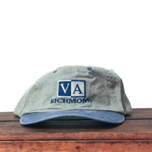 May include: A gray baseball cap with a blue brim. The cap features the letters "VA" in a blue and white square, with "RICHMOND" embroidered below in blue. The hat is resting on a wooden surface.