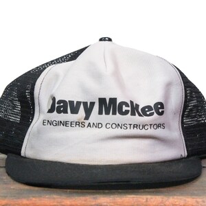 Puede incluir: Una gorra de camionero blanca y negra con el texto "Javy McKee Engineers and Constructors" impreso en la parte delantera.