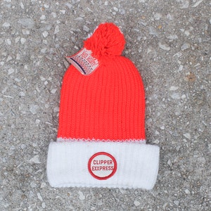 Puede incluir: Un gorro de punto naranja neón con un borde blanco y un pompón a juego. El borde blanco presenta un parche bordado en rojo y blanco que dice "CLIPPER EXXPRESS". Una etiqueta que dice "Yea Team!" está adjunta.