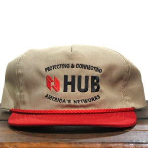 May include: Tan and red baseball cap with the text "PROTECTING & CONNECTING HUB AMERICA'S NETWORKS". The cap has a red brim and a red rope detail. The hat is made of a durable fabric.