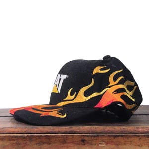 Puede incluir: Gorra de b&eacute;isbol negra con el logotipo CAT en blanco y amarillo. La gorra presenta un dise&ntilde;o de llamas en amarillo, naranja y rojo. La visera tambi&eacute;n es negra con el dise&ntilde;o de llamas.