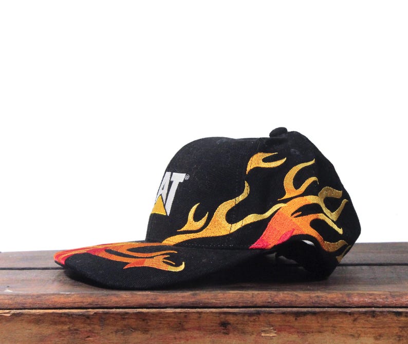 Puede incluir: Gorra de b&eacute;isbol negra con el logotipo CAT en blanco y amarillo. La gorra presenta un dise&ntilde;o de llamas en amarillo, naranja y rojo. La visera tambi&eacute;n es negra con el dise&ntilde;o de llamas.