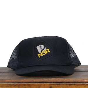 May include: Black trucker cap with a mesh back and a curved peak. The front displays a yellow and white NSR logo patch. The cap is on a wooden surface.