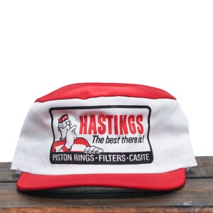 May include: A red and white baseball cap with a logo for Hastings Piston Rings, Filters, and Casite. The logo features a cartoon man with a finger pointing to the text "Hastings The best there is!"