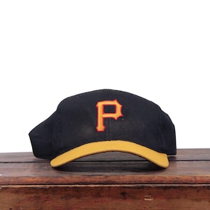 May include: A black baseball cap featuring a prominent letter P embroidered on the front in yellow with a red outline. The cap has a solid yellow brim and a structured crown, presenting a classic athletic design with bold, contrasting colours.