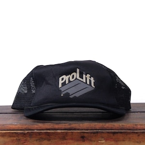 May include: Black trucker hat with the text "ProLift" in white and gray. The hat has a mesh back and a curved brim. The hat is sitting on a wooden surface.