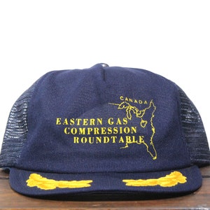 May include: A navy blue trucker hat with a black mesh back. The hat has a yellow embroidered logo that says "Eastern Gas Compression Roundtable" and a map of Canada. The hat also has a yellow embroidered wing on each side.
