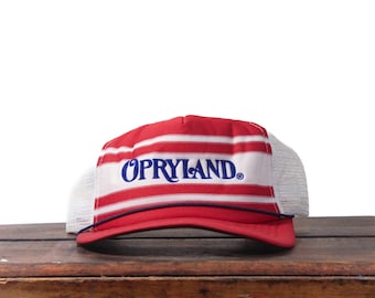 May include: Red and white striped trucker hat with the word "OPRYLAND" in blue script across the front. The hat has a white mesh back and a navy blue rope detail along the brim. A vintage-style cap.