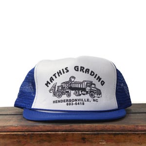 Peut inclure: Casquette de camionneur bleue et blanche avec le texte "MATHIS GRADING" et une illustration de véhicules de chantier. La casquette a une visière bleue et un dos en filet, avec la ville et le numéro de téléphone imprimés en dessous.