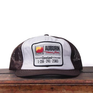 以下が含まれることがあります： 「AUBURN」と「Versa-Hoe」の文字が入った長方形のロゴが特徴の白いフロントパネルが付いたトラッカーハット。茶色のつばとメッシュのバック、電話番号1-216-241-2383が特徴です。